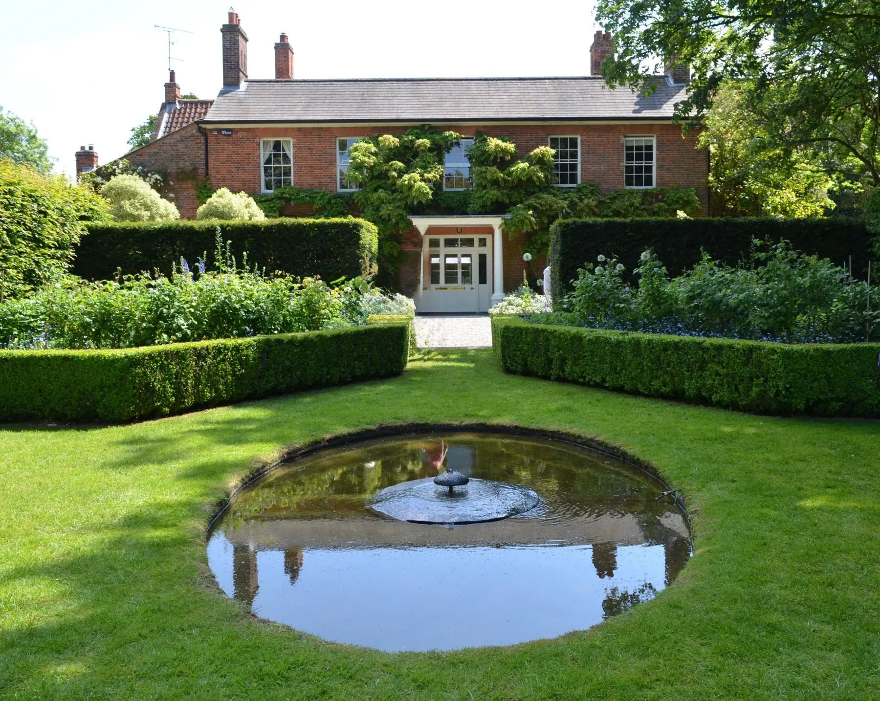 Formal English garden with a round pond and neat hedges in front of a brick house